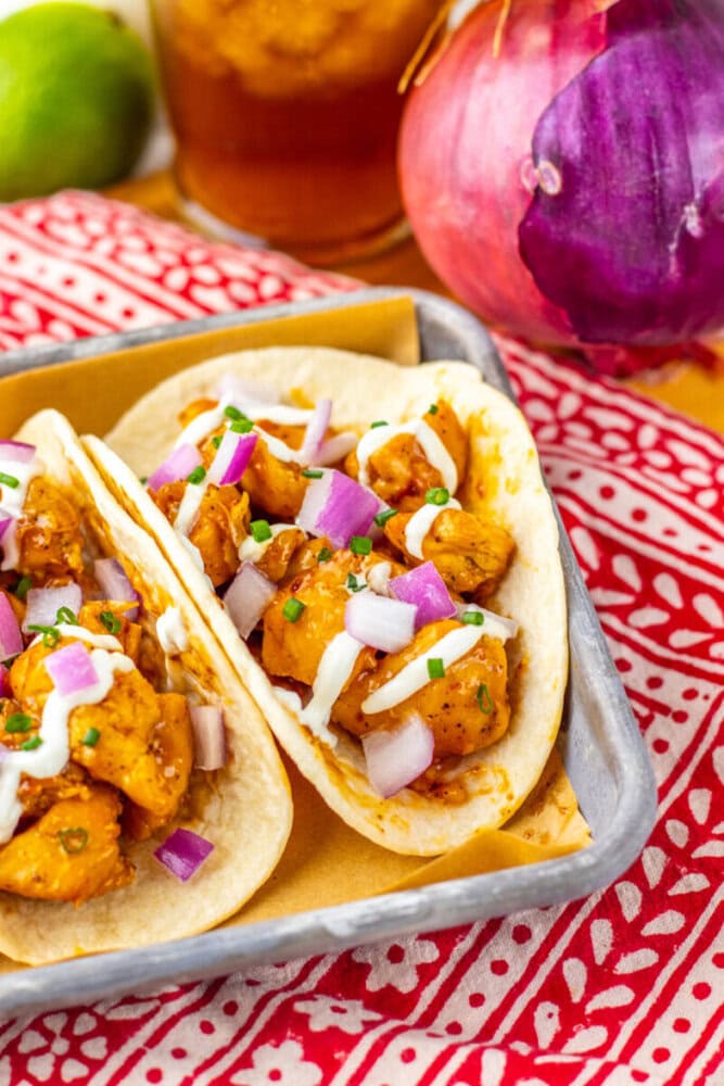 Two soft tacos filled with seasoned chicken, diced red onions, chopped herbs, and drizzled with white sauce, served on a tray with a red patterned cloth and vegetables in the background.