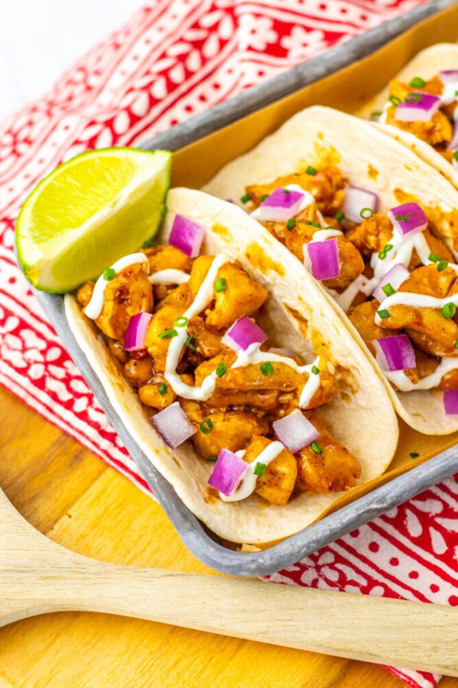 Two chicken tacos in soft tortillas topped with diced red onion, creamy sauce, and chopped herbs, served with a lime wedge on a patterned red cloth.