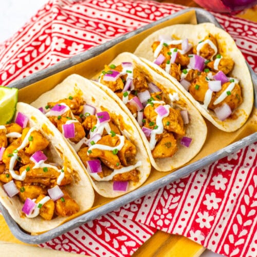 Four chicken tacos topped with diced red onions, cilantro, and a drizzle of white sauce, served on a tray with a lime wedge on a red patterned cloth.