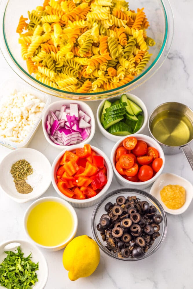 A variety of ingredients for pasta salad are arranged on a counter, including tri-color rotini, chopped vegetables, black olives, cheese, seasonings, oil, and a lemon.