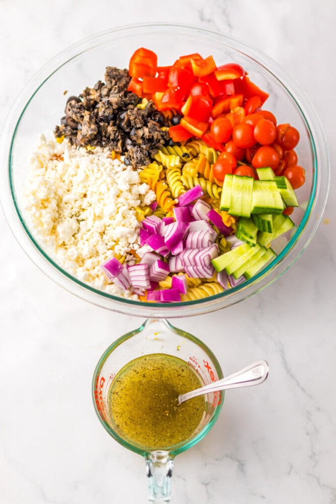 A glass bowl with pasta, feta cheese, chopped black olives, red onion, cucumber, cherry tomatoes, and red bell pepper beside a measuring cup of vinaigrette dressing with a spoon.