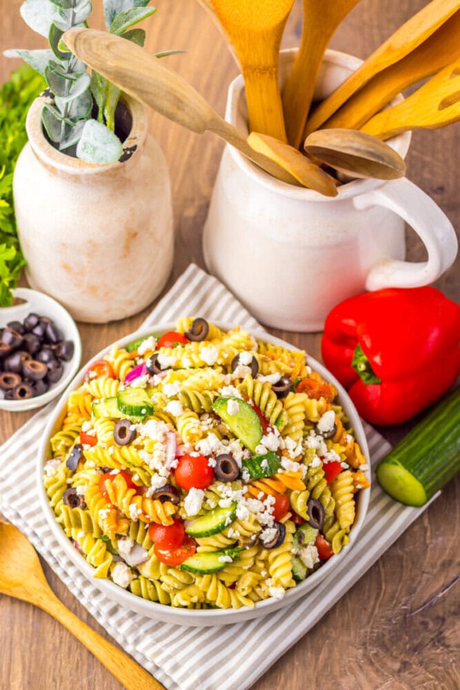 A bowl of pasta salad with cherry tomatoes, cucumbers, olives, and feta cheese sits on a striped cloth next to fresh vegetables, a bowl of olives, and wooden utensils in ceramic containers.