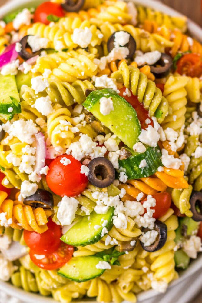 A close-up of a pasta salad with rotini noodles, cherry tomatoes, cucumber slices, black olives, red onion, and crumbled feta cheese.