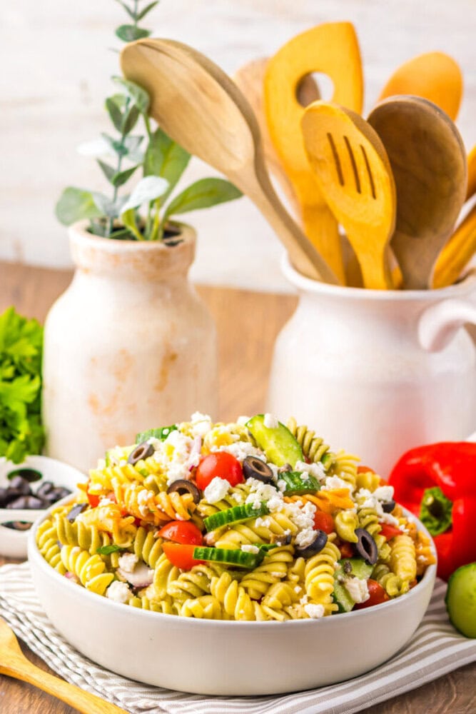 A bowl of pasta salad with vegetables and feta cheese sits on a table, with kitchen utensils and a potted plant in the background.