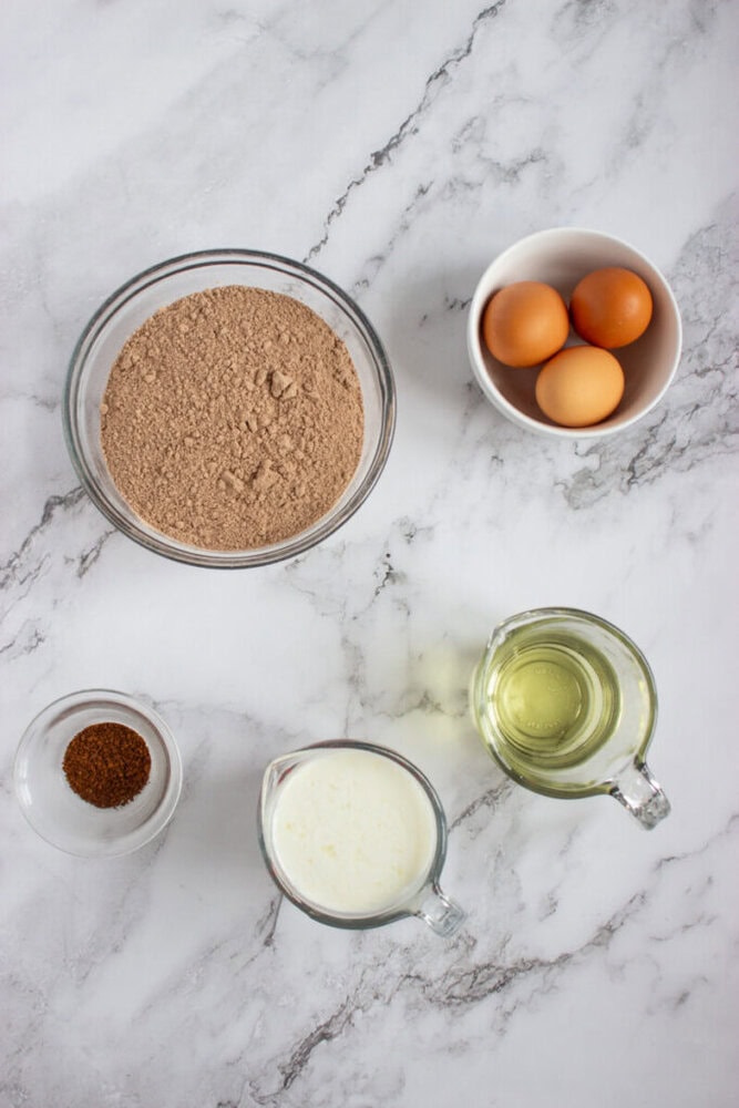 Five ingredients for baking are arranged on a marble surface: a bowl of eggs, a bowl of cocoa powder mix, a small bowl of instant coffee, a cup of milk, and a cup of oil.
