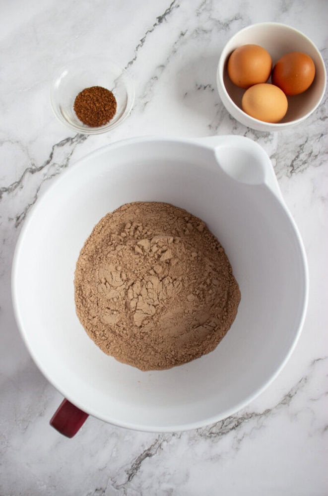 A large white mixing bowl with cocoa powder and flour sits on a marble surface, next to a small bowl of brown sugar and a bowl containing three eggs.