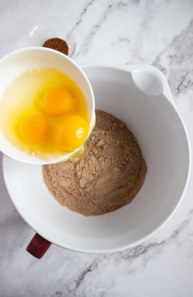 A bowl with cocoa powder and another bowl with three raw eggs being poured in, on a marble countertop.