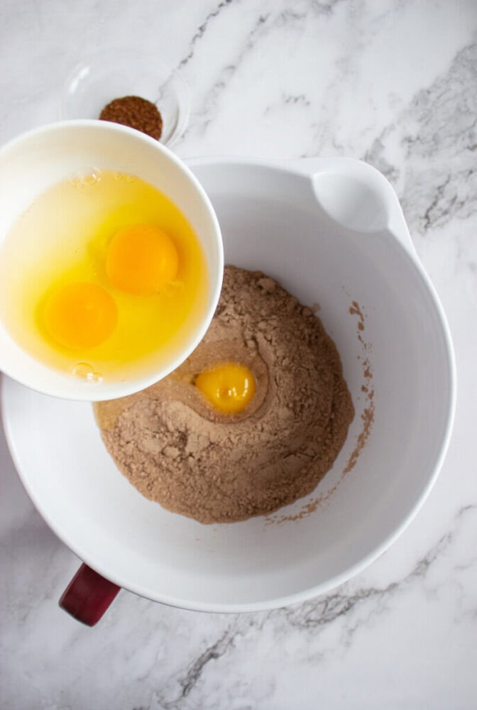 Three raw eggs in a bowl being poured into a mixing bowl with chocolate cake mix, on a marble countertop.