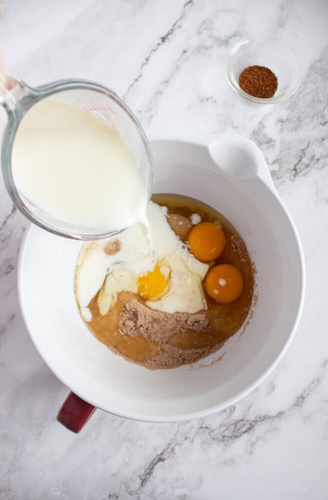 Milk is being poured from a measuring cup into a mixing bowl containing eggs, flour, sugar, and other ingredients, with a small bowl of spices nearby.