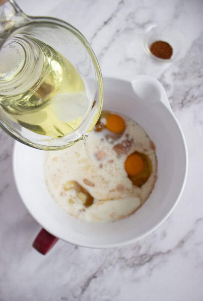 A clear glass jug pours oil into a mixing bowl containing eggs, milk, and dry ingredients. A small bowl of spices sits nearby on a marble countertop.