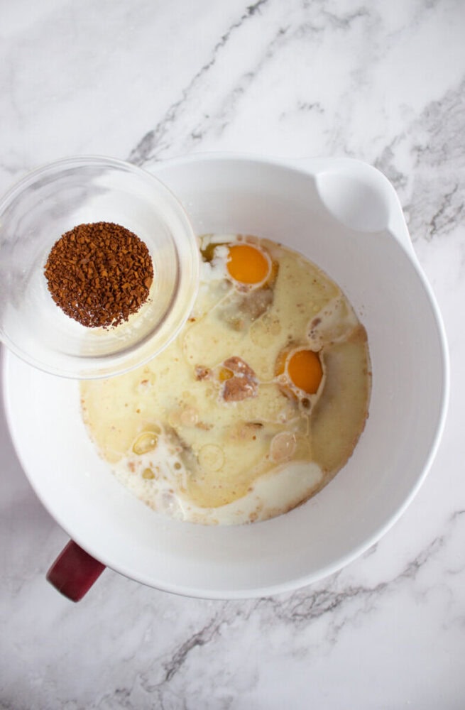 A mixing bowl with eggs, milk, and other wet ingredients, with a small bowl of instant coffee granules being held above it on a marble countertop.