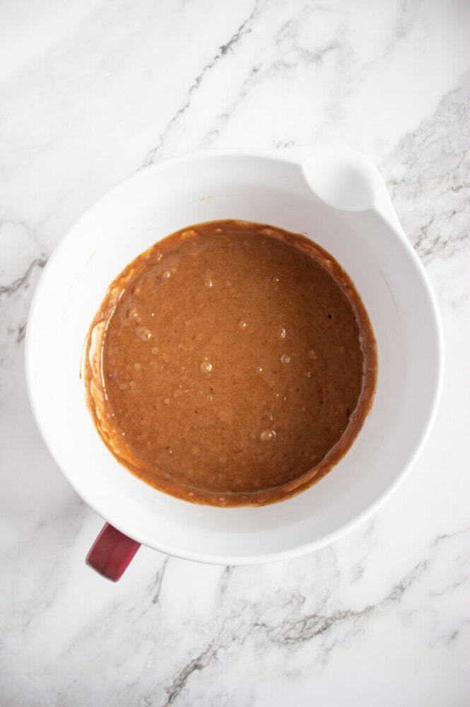 A white mixing bowl containing a light brown batter sits on a marble countertop.