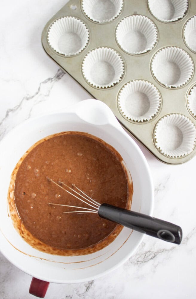A mixing bowl with chocolate batter and a whisk sits next to a muffin tin lined with paper baking cups on a marble countertop.
