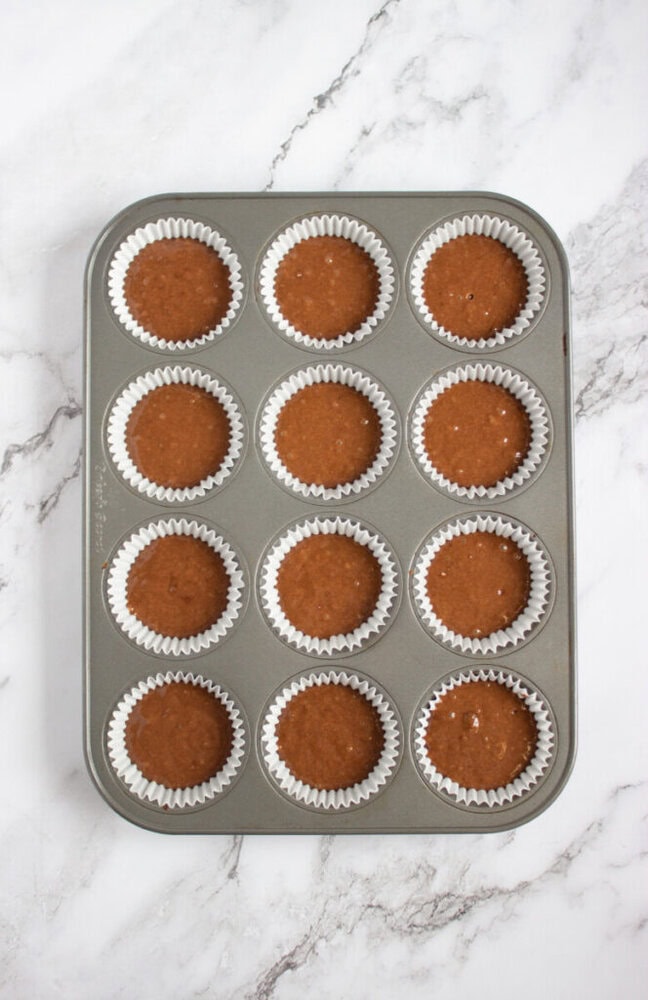 A muffin tin with twelve paper liners, each filled with chocolate batter, sits on a white marble surface.