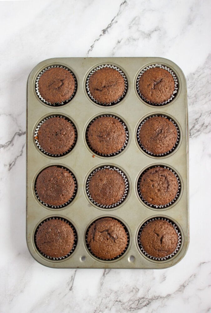 A metal muffin tin filled with twelve baked chocolate cupcakes in paper liners, placed on a white marble surface.