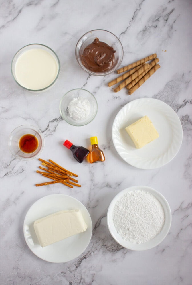Assorted baking ingredients including cream, Nutella, butter, cream cheese, powdered sugar, pretzel sticks, wafer rolls, vanilla, and food coloring on a marble surface.