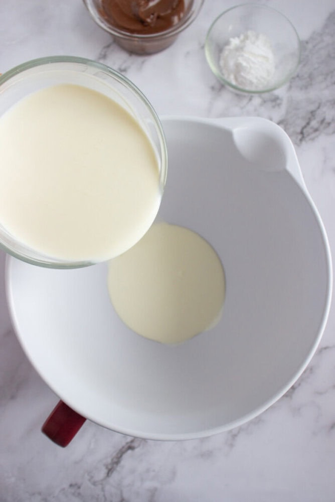 A glass bowl of cream is being poured into a white mixing bowl on a marble surface, with small bowls of chocolate and powdered sugar nearby.