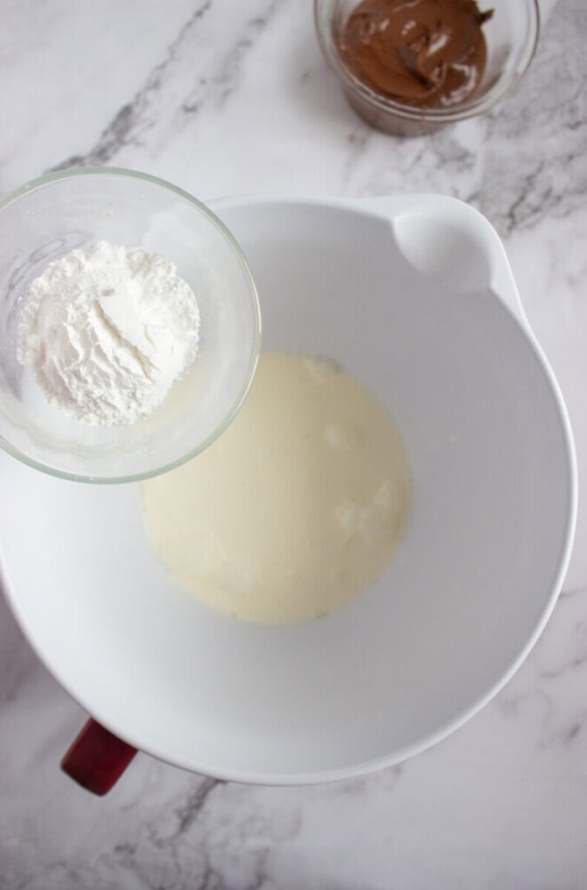 A glass bowl with flour is held above a white mixing bowl containing a liquid mixture, with a small bowl of chocolate nearby on a marble countertop.