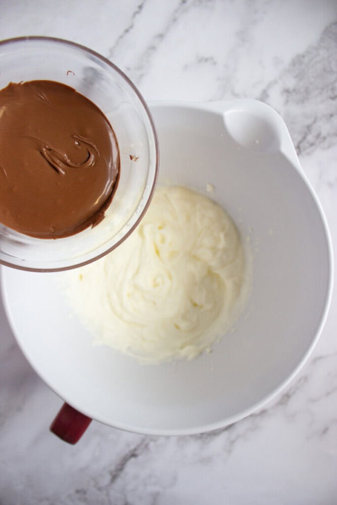 A bowl of melted chocolate is held above a larger mixing bowl containing a creamy white mixture on a marble countertop.