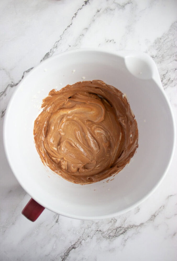 A white mixing bowl with light brown batter swirled inside, sitting on a white marble countertop.