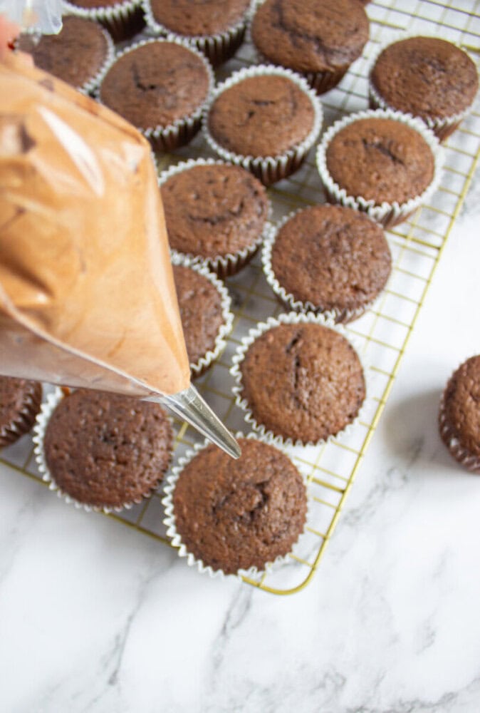 A hand piping chocolate frosting onto a chocolate cupcake, with more cupcakes cooling on a wire rack in the background.