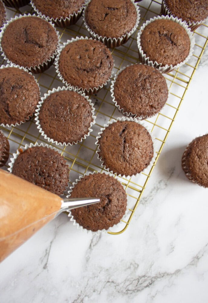 A piping bag filled with chocolate frosting is positioned over chocolate cupcakes cooling on a wire rack, set on a marble surface.