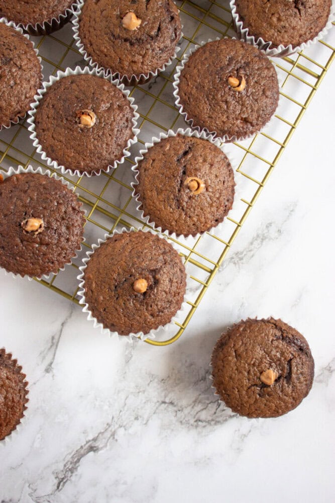 Chocolate muffins with chocolate chips, each in a white paper liner, are cooling on a wire rack on a marble surface. One muffin is placed off the rack.