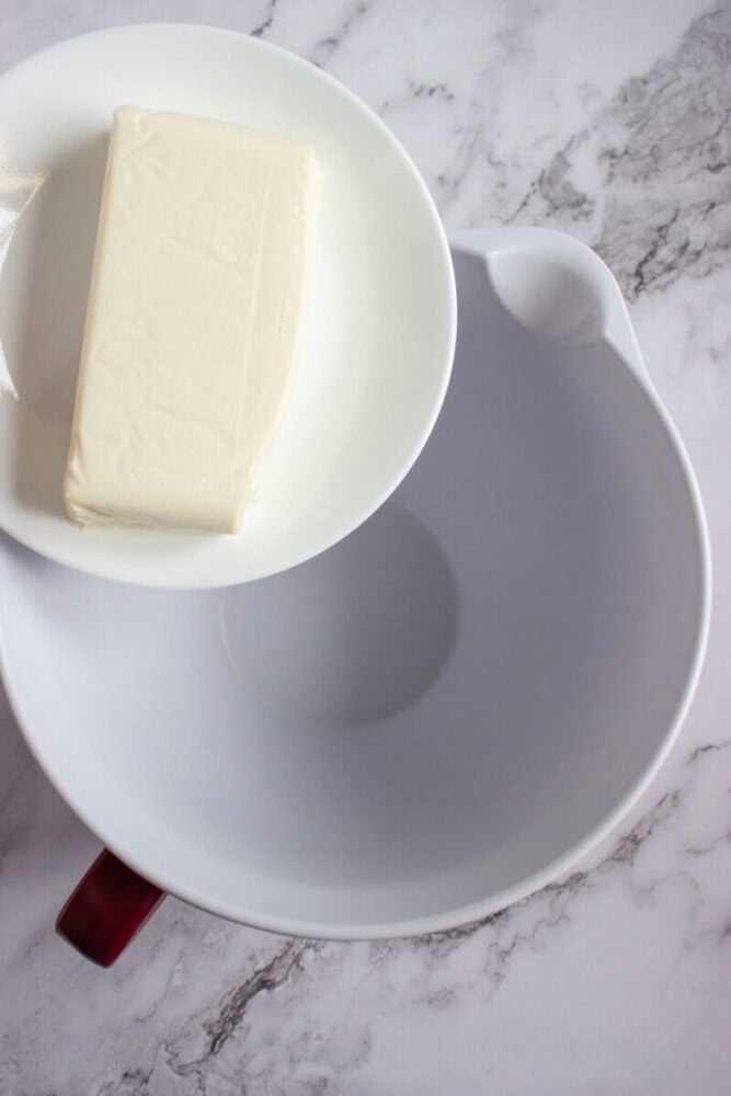 A white plate holding a block of cream cheese is positioned above a large empty mixing bowl on a marble countertop.