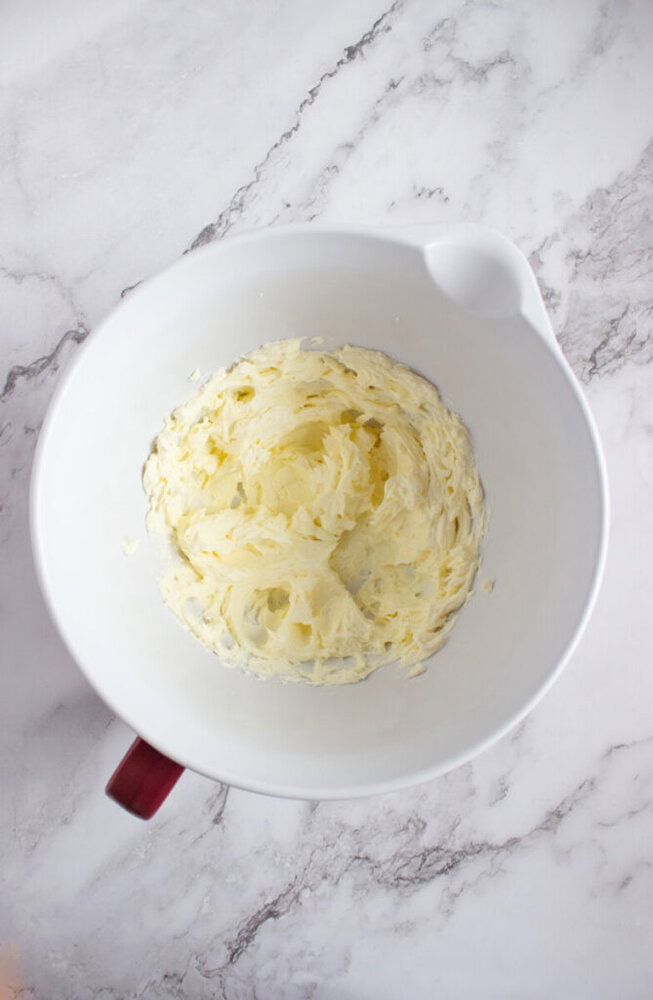 A white mixing bowl with whipped, pale yellow butter mixture sits on a marble countertop.
