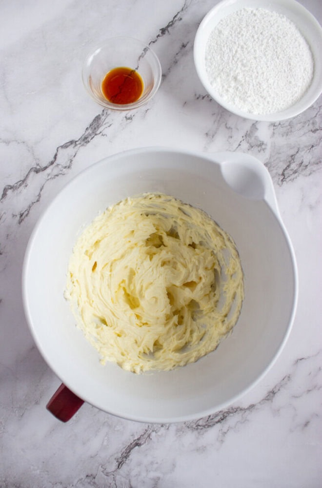 A mixing bowl with creamed butter, a small bowl of vanilla extract, and a bowl of powdered sugar on a marble countertop.
