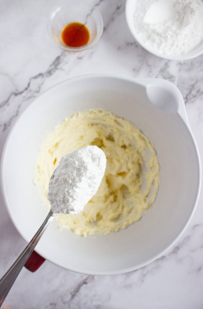 A spoonful of powdered sugar is held above a mixing bowl with creamed butter, with a bowl of vanilla extract and a bowl of flour nearby on a marble surface.