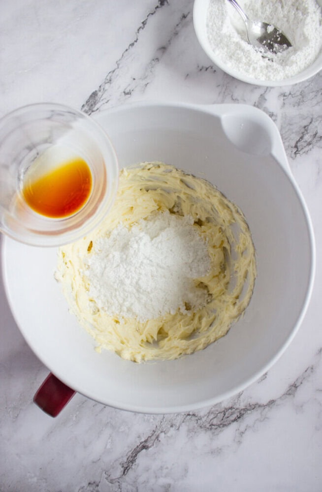 A mixing bowl with creamed butter and powdered sugar, with a small cup of vanilla extract being poured in. A bowl of powdered sugar and a spoon are in the background.