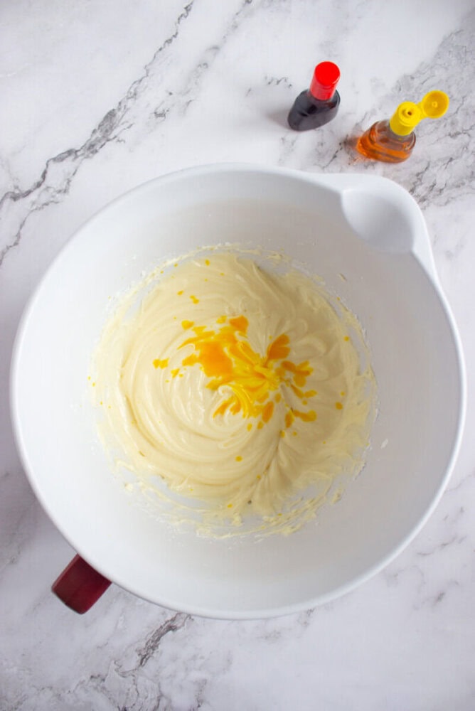 A white mixing bowl with creamy batter and yellow food coloring drops, next to bottles of red and yellow coloring on a marble countertop.