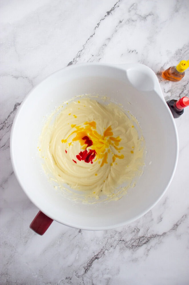 A white mixing bowl with creamy batter and red and yellow food coloring drops, on a marble countertop with food coloring bottles nearby.