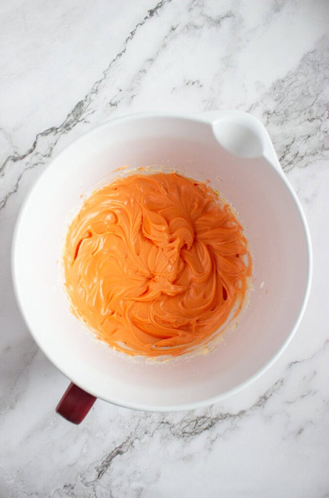 A white mixing bowl with orange frosting swirled inside sits on a white marble countertop.