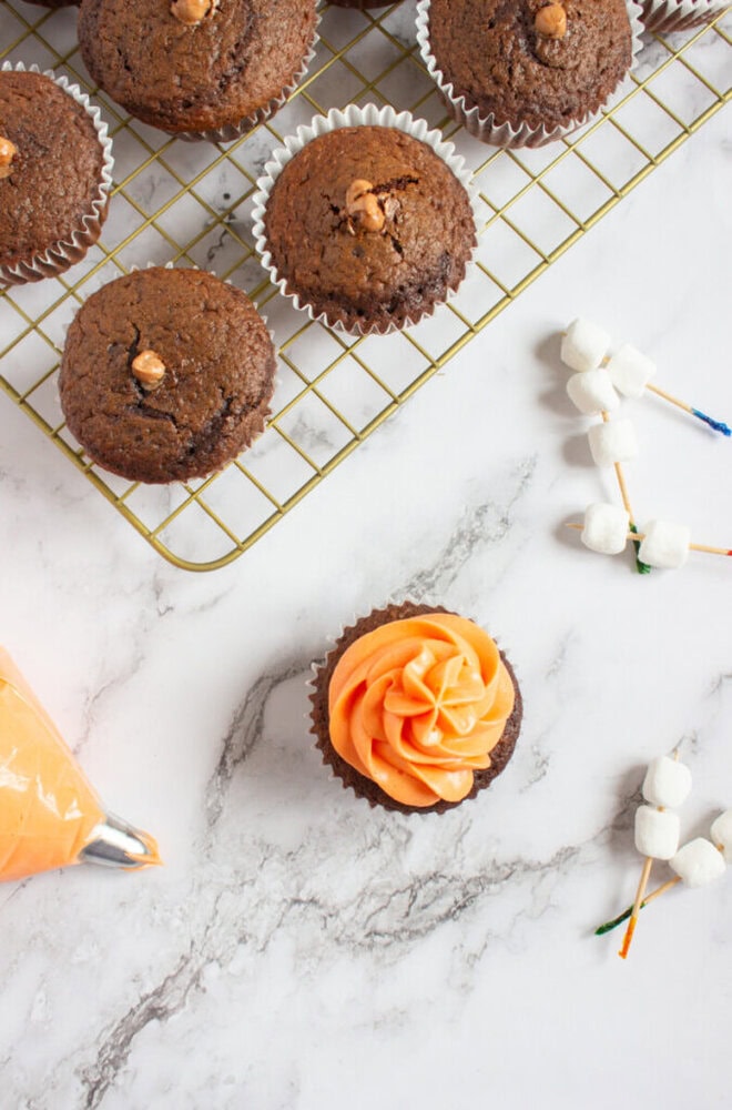 Chocolate cupcakes on a cooling rack, one cupcake with orange frosting on a marble surface, piping bag, and marshmallows on toothpicks nearby.