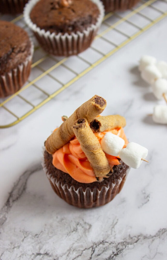 A chocolate cupcake with orange frosting, topped with rolled wafer cookies and two mini marshmallows on a toothpick, sits on a marble surface near a cooling rack.