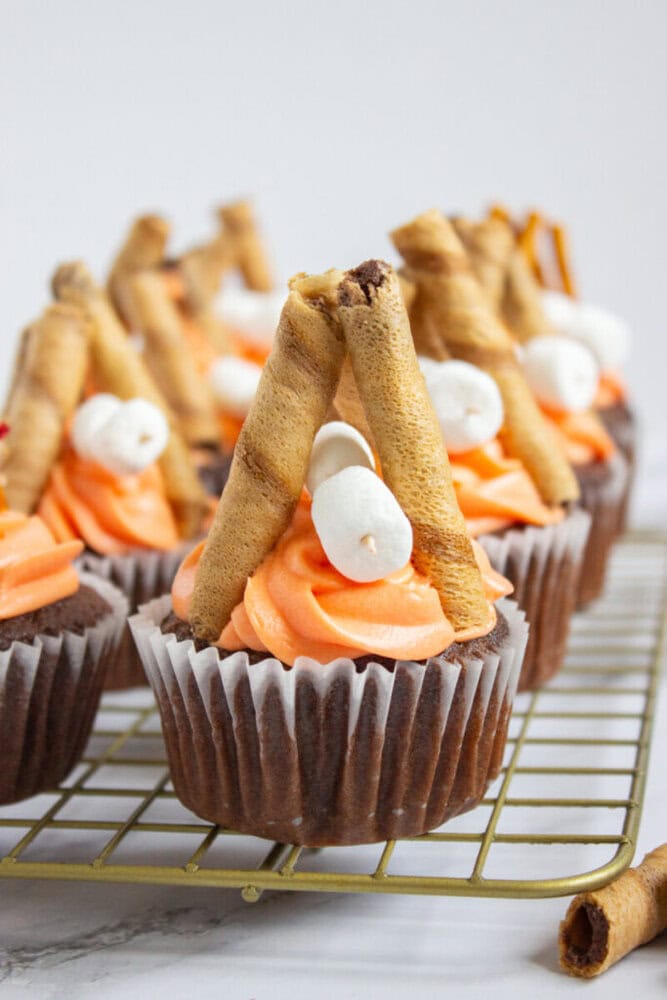 Chocolate cupcakes with orange frosting are topped with rolled wafer cookies arranged like teepees and small white marshmallows, displayed on a cooling rack.