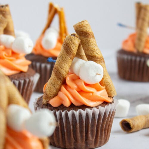 Chocolate cupcakes topped with orange frosting, wafer rolls arranged like campfires, and mini marshmallows as decoration.
