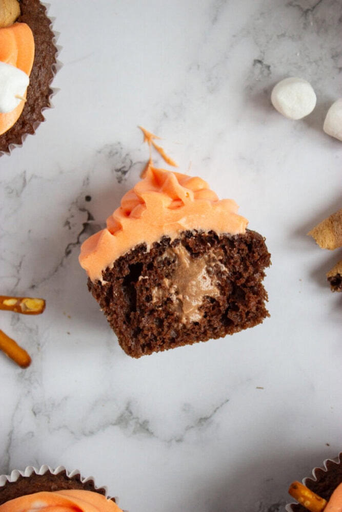 A chocolate cupcake with orange frosting is cut in half, revealing a creamy filling inside. Surrounding the cupcake are mini marshmallows, pretzel sticks, and other dessert items.