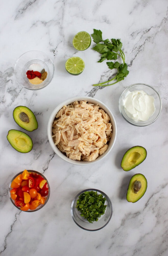 A bowl of shredded chicken surrounded by avocado halves, chopped tomatoes, sour cream, spices, lime halves, chopped cilantro, and fresh parsley on a marble surface.