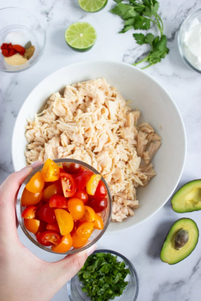 A hand holds a bowl of halved cherry tomatoes above a larger bowl of shredded chicken, surrounded by avocado, lime, cilantro, spices, and sour cream on a marble countertop.