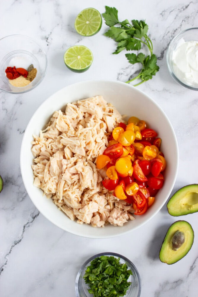 A white bowl with shredded chicken and halved cherry tomatoes sits on a marble surface, surrounded by avocado, lime, cilantro, spices, and a bowl of sour cream.