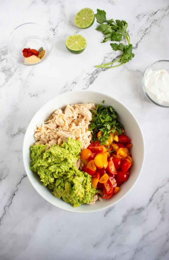 A white bowl contains shredded chicken, mashed avocado, chopped cilantro, and halved cherry tomatoes. Nearby are a sliced lime, spices, cilantro, and a bowl of sour cream.