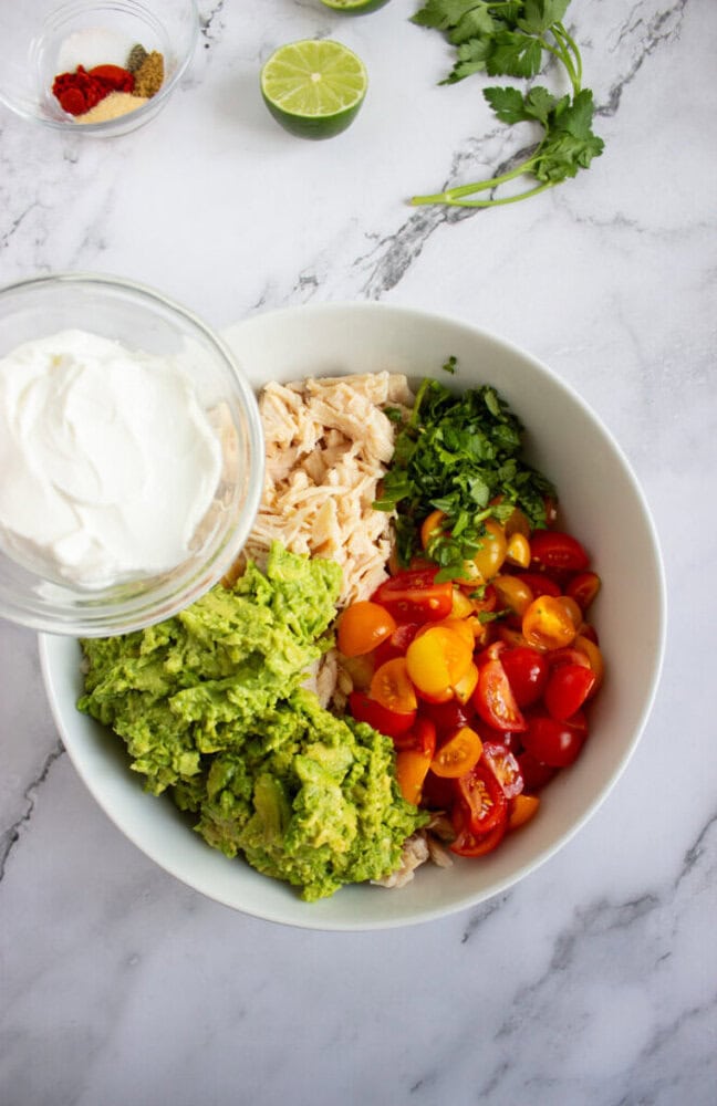 A white bowl with shredded chicken, chopped cilantro, halved cherry tomatoes, guacamole, and a separate bowl of sour cream on a marble surface with assorted ingredients nearby.