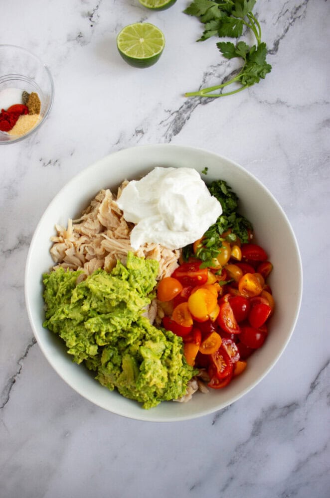 A bowl containing shredded chicken, mashed avocado, halved cherry tomatoes, chopped cilantro, and a dollop of sour cream, with spices, lime, and herbs nearby on a marble surface.