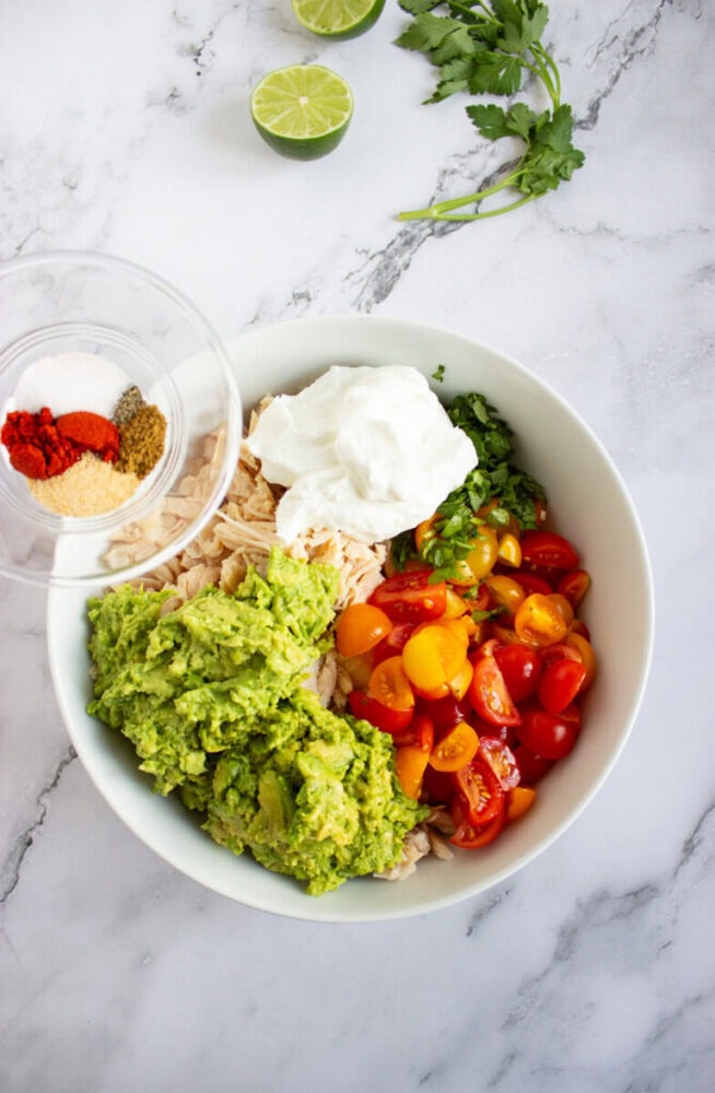 A white bowl contains mashed avocado, halved cherry tomatoes, chopped cilantro, shredded chicken, and a dollop of sour cream. Nearby are lime halves, parsley, and a small bowl of spices.