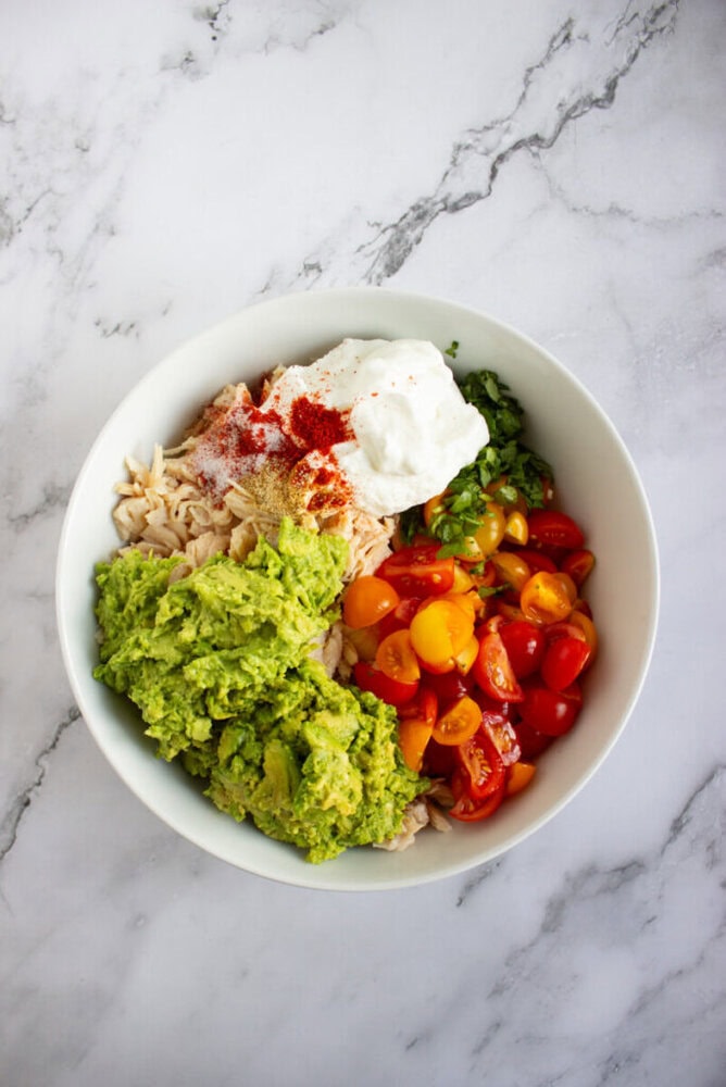 A white bowl containing mashed avocado, sliced cherry tomatoes, shredded chicken, cooked rice, cilantro, Greek yogurt, and a sprinkle of paprika on a marble surface.