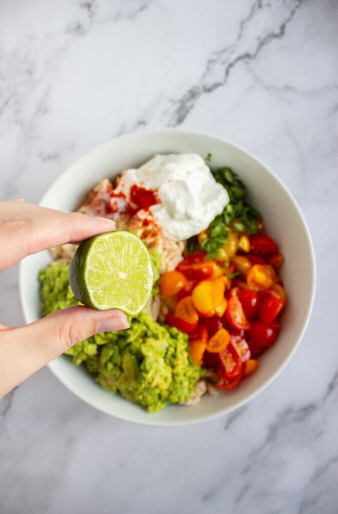A hand holds half a lime above a bowl filled with guacamole, chopped tomatoes, rice, herbs, and a dollop of sour cream on a marble surface.
