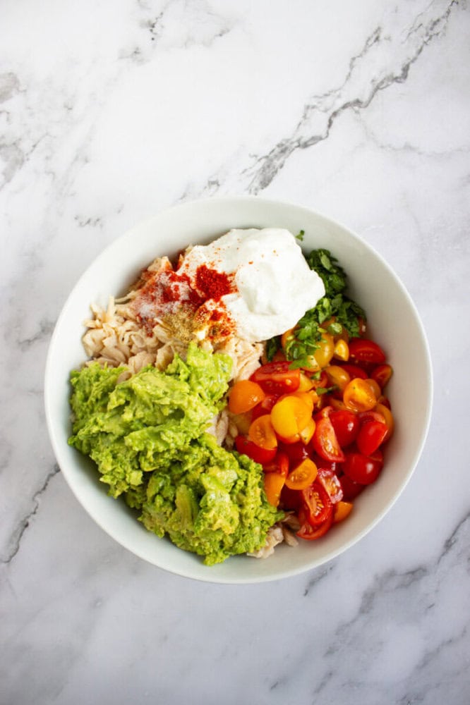 A white bowl containing shredded chicken, mashed avocado, halved cherry tomatoes, chopped herbs, a dollop of yogurt, and sprinkled spices on a marble surface.